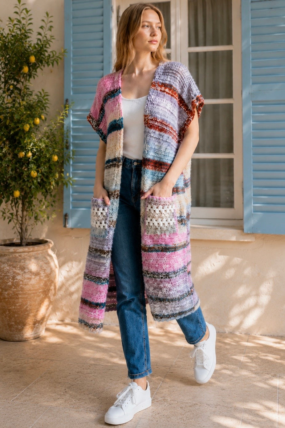 A woman wearing a long, open-front, short-sleeved chunky knit cardigan with horizontal multicolor stripes in pink, blue, beige, brown, and white, paired with a white tank top and blue jeans, standing in front of a window with blue shutters.