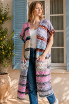 A woman wearing a long, open-front, short-sleeved chunky knit cardigan with horizontal multicolor stripes in pink, blue, beige, brown, and white, paired with a white tank top and blue jeans, standing in front of a window with blue shutters.