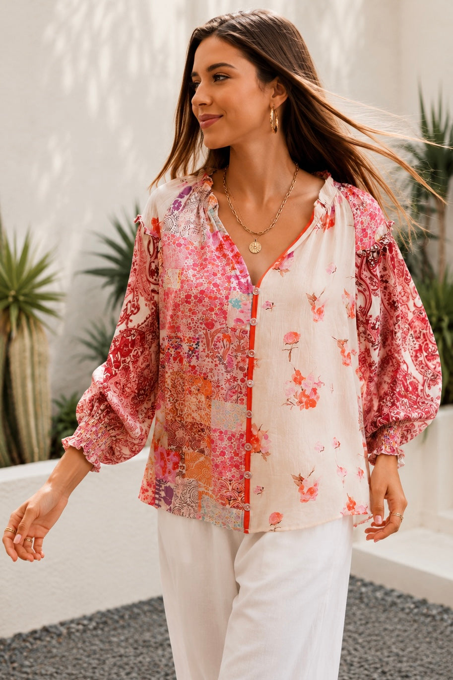 A woman wearing a bohemian-style long-sleeve blouse with a pink, red, and orange floral patchwork print on a white base, styled with white pants and gold jewelry in an outdoor setting.