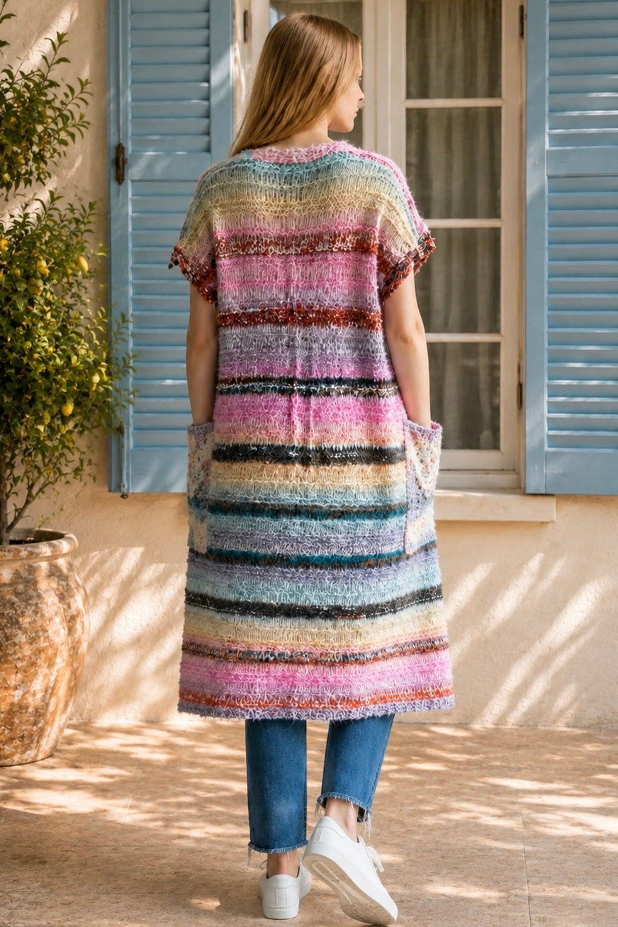 A woman wearing a long, open-front, short-sleeved chunky knit cardigan with horizontal multicolor stripes in pink, blue, beige, brown, and white, paired with a white tank top and blue jeans, standing in front of a window with blue shutters.