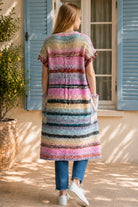 A woman wearing a long, open-front, short-sleeved chunky knit cardigan with horizontal multicolor stripes in pink, blue, beige, brown, and white, paired with a white tank top and blue jeans, standing in front of a window with blue shutters.