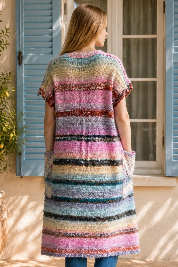 A woman wearing a long, open-front, short-sleeved chunky knit cardigan with horizontal multicolor stripes in pink, blue, beige, brown, and white, paired with a white tank top and blue jeans, standing in front of a window with blue shutters.