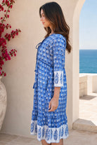 A woman wearing a blue and white patterned cotton tunic dress with tie details and a ruffled hem, standing in a Mediterranean-style setting with a sea view in the background from Bohemian Love Runway.