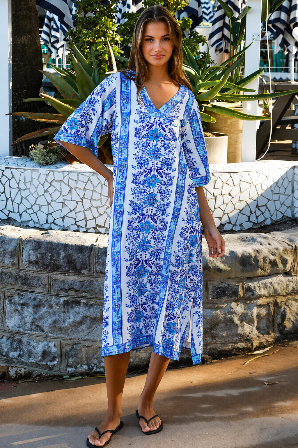 A woman wearing a blue and white patterned kaftan dress with a V-neck and three-quarter sleeves, walking outdoors in front of a stone wall and tropical plants.