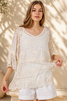 A woman wearing a cream lace top with three-quarter sleeves and white shorts, standing in front of a warm beige wall with leaf shadows.