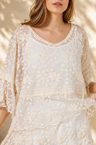 A woman wearing a cream lace top with three-quarter sleeves and white shorts, standing in front of a warm beige wall with leaf shadows.