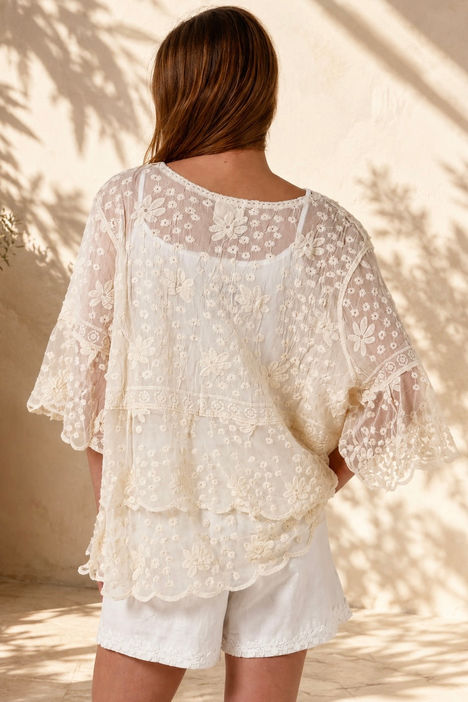 A woman wearing a cream lace top with three-quarter sleeves and white shorts, standing in front of a warm beige wall with leaf shadows.