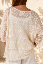 A woman wearing a cream lace top with three-quarter sleeves and white shorts, standing in front of a warm beige wall with leaf shadows.