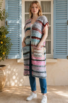 A woman wearing a long, open-front, short-sleeved chunky knit cardigan with horizontal multicolor stripes in pink, blue, beige, brown, and white, paired with a white tank top and blue jeans, standing in front of a window with blue shutters.