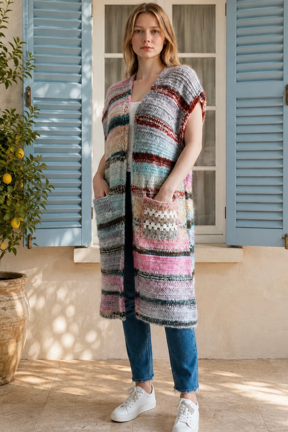 A woman wearing a long, open-front, short-sleeved chunky knit cardigan with horizontal multicolor stripes in pink, blue, beige, brown, and white, paired with a white tank top and blue jeans, standing in front of a window with blue shutters.