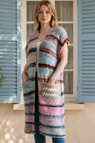 A woman wearing a long, open-front, short-sleeved chunky knit cardigan with horizontal multicolor stripes in pink, blue, beige, brown, and white, paired with a white tank top and blue jeans, standing in front of a window with blue shutters.