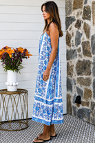 A woman modeling a blue and white patterned maxi dress with spaghetti straps, standing on a patterned tile porch next to a small table with orange flowers.