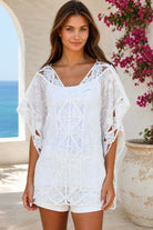 A woman wearing a white eyelet lace cover-up top with cutout details, standing outdoors in front of a white stucco wall with a blue ocean and pink flowers in the background.