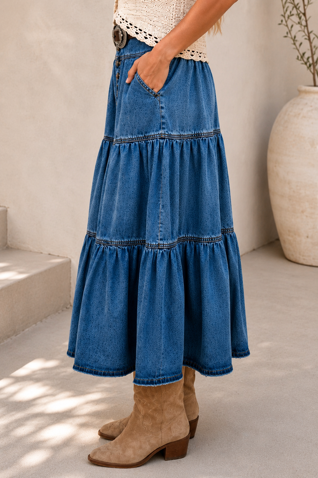 A woman models a bohemian-style outfit featuring a high-waisted, tiered blue denim skirt with a decorative belt, paired with a cream crochet top and beige boots, set against a sunlit neutral background.