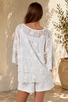 A woman wearing a white lace long-sleeve tunic top with matching white shorts, standing in a sunlit outdoor setting with a beige wall and potted plant in the background.