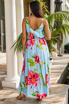 A woman viewed from behind wearing a light blue sleeveless maxi dress with a vibrant pink and green floral print, standing on a stone patio in front of white columns and palm trees.