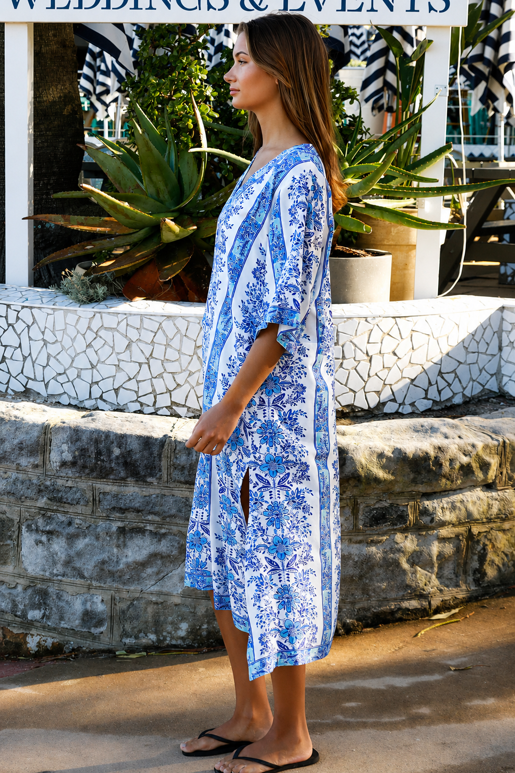 A woman wearing a blue and white patterned kaftan dress with a V-neck and three-quarter sleeves, walking outdoors in front of a stone wall and tropical plants.