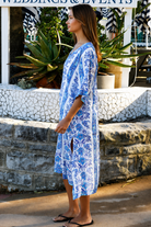 A woman wearing a blue and white patterned kaftan dress with a V-neck and three-quarter sleeves, walking outdoors in front of a stone wall and tropical plants.