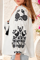 A woman wearing a white long-sleeve blouse with black floral and bird embroidery, paired with light blue jeans, photographed against a textured stucco wall with flowers in the background. Bohemian Love Runway.