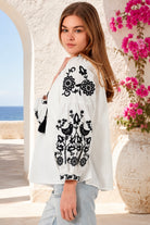 A woman wearing a white long-sleeve blouse with black floral and bird embroidery, paired with light blue jeans, standing in a sunlit outdoor setting with a white archway, blue sea in the background, and pink flowers on the right from Bohemian Love Runway.