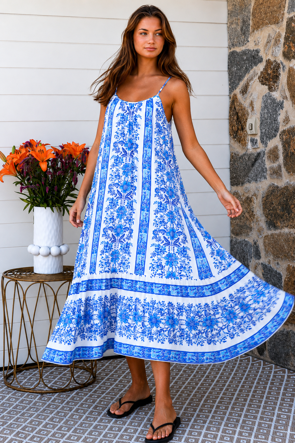 A woman modeling a blue and white patterned maxi dress with spaghetti straps, standing on a patterned tile porch next to a small table with orange flowers.