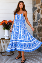 A woman modeling a blue and white patterned maxi dress with spaghetti straps, standing on a patterned tile porch next to a small table with orange flowers.