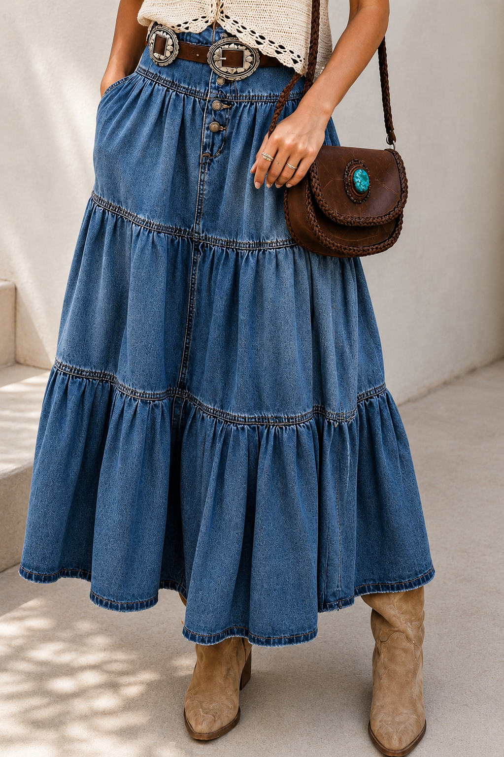 A woman models a bohemian-style outfit featuring a high-waisted, tiered blue denim skirt with a decorative belt, paired with a cream crochet top and beige boots, set against a sunlit neutral background.