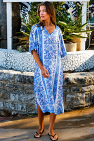A woman wearing a blue and white patterned kaftan dress with a V-neck and three-quarter sleeves, walking outdoors in front of a stone wall and tropical plants.