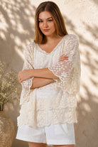 A woman wearing a cream lace top with three-quarter sleeves and white shorts, standing in front of a warm beige wall with leaf shadows.
