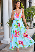 A woman wearing a light blue sleeveless maxi dress with a vibrant pink and green floral print, standing outdoors in front of white columns and tropical plants.