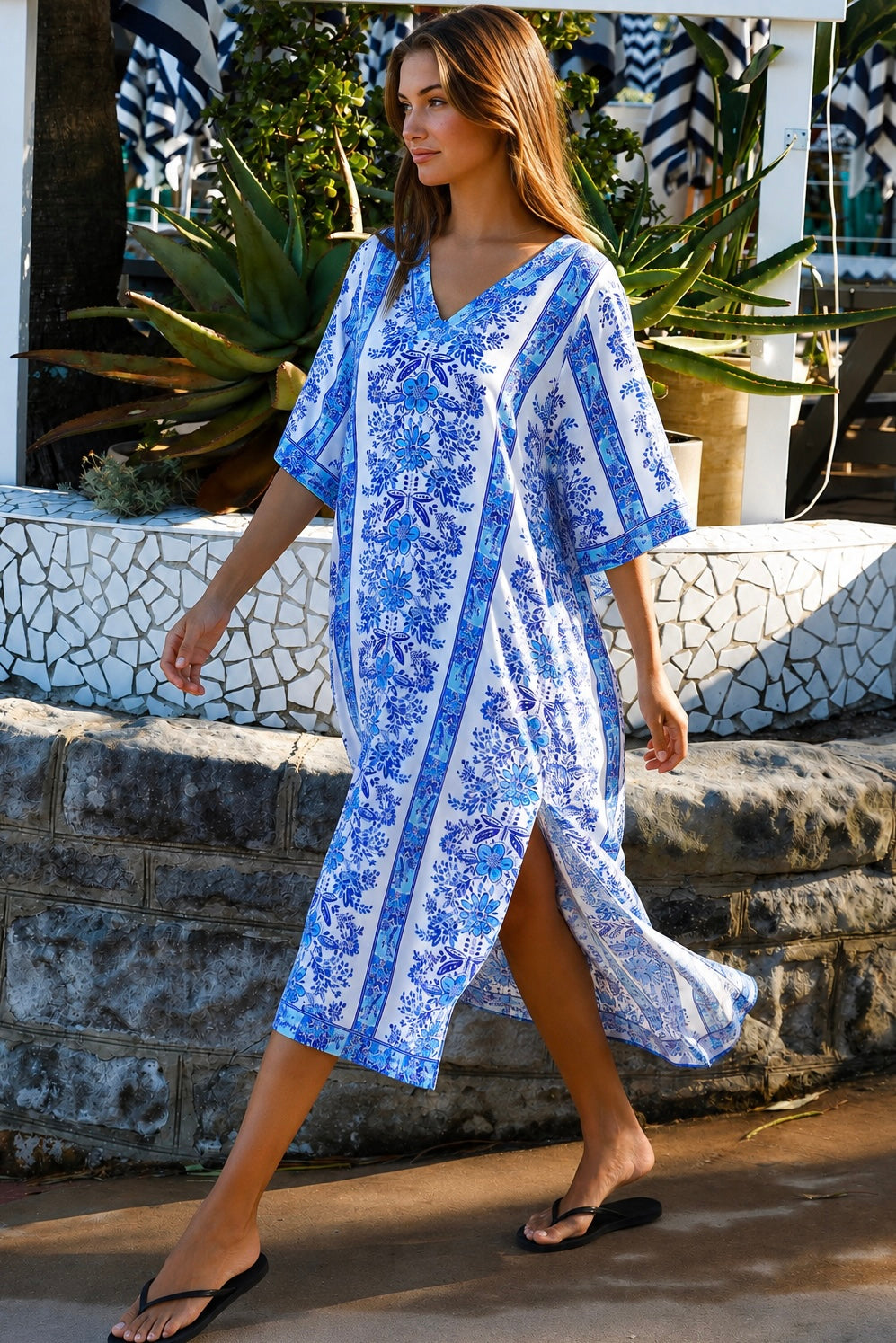 A woman wearing a blue and white patterned kaftan dress with a V-neck and three-quarter sleeves, walking outdoors in front of a stone wall and tropical plants.