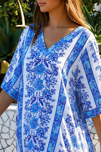 A woman wearing a white kaftan-style tunic dress with a vibrant blue floral and geometric pattern, photographed outdoors with green foliage in the background.