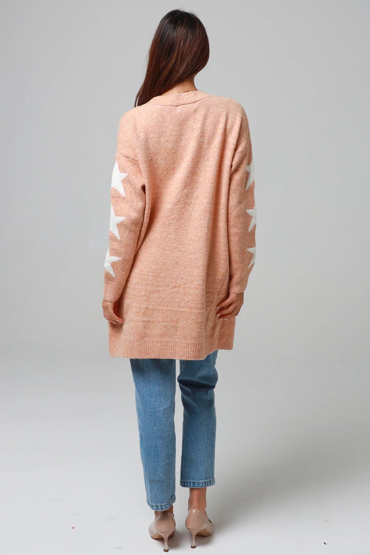 Woman wearing a peachy-blush longline cardigan with white star accents, front pockets, and relaxed fit — cozy knitwear styled with jeans and a white top, perfect for layering.