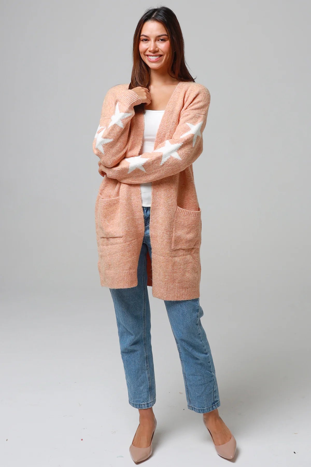 Woman wearing a peachy-blush longline cardigan with white star accents, front pockets, and relaxed fit — cozy knitwear styled with jeans and a white top, perfect for layering.