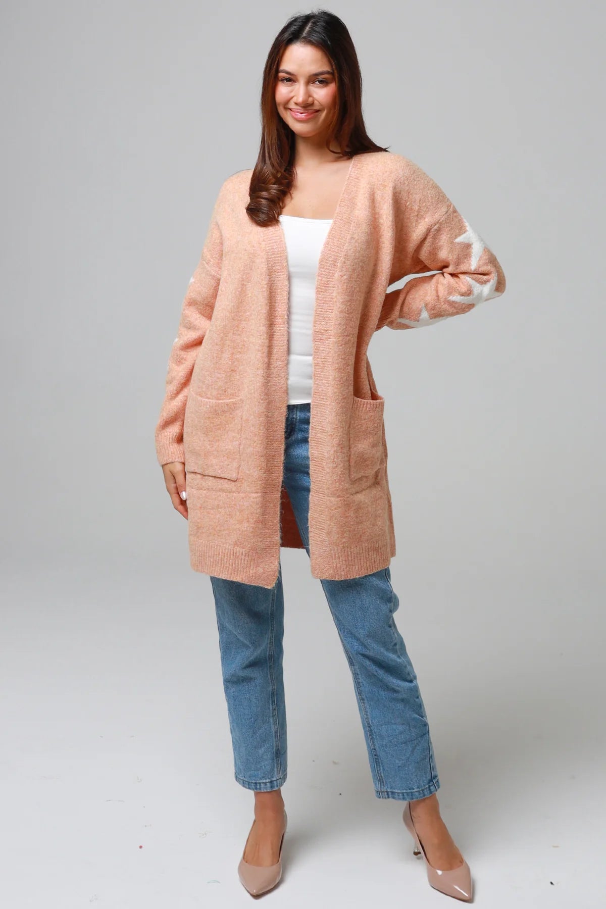 Woman wearing a peachy-blush longline cardigan with white star accents, front pockets, and relaxed fit — cozy knitwear styled with jeans and a white top, perfect for layering.