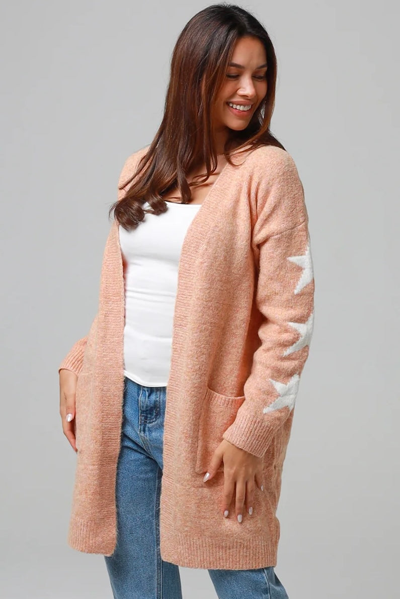 Woman wearing a peachy-blush longline cardigan with white star accents, front pockets, and relaxed fit — cozy knitwear styled with jeans and a white top, perfect for layering.