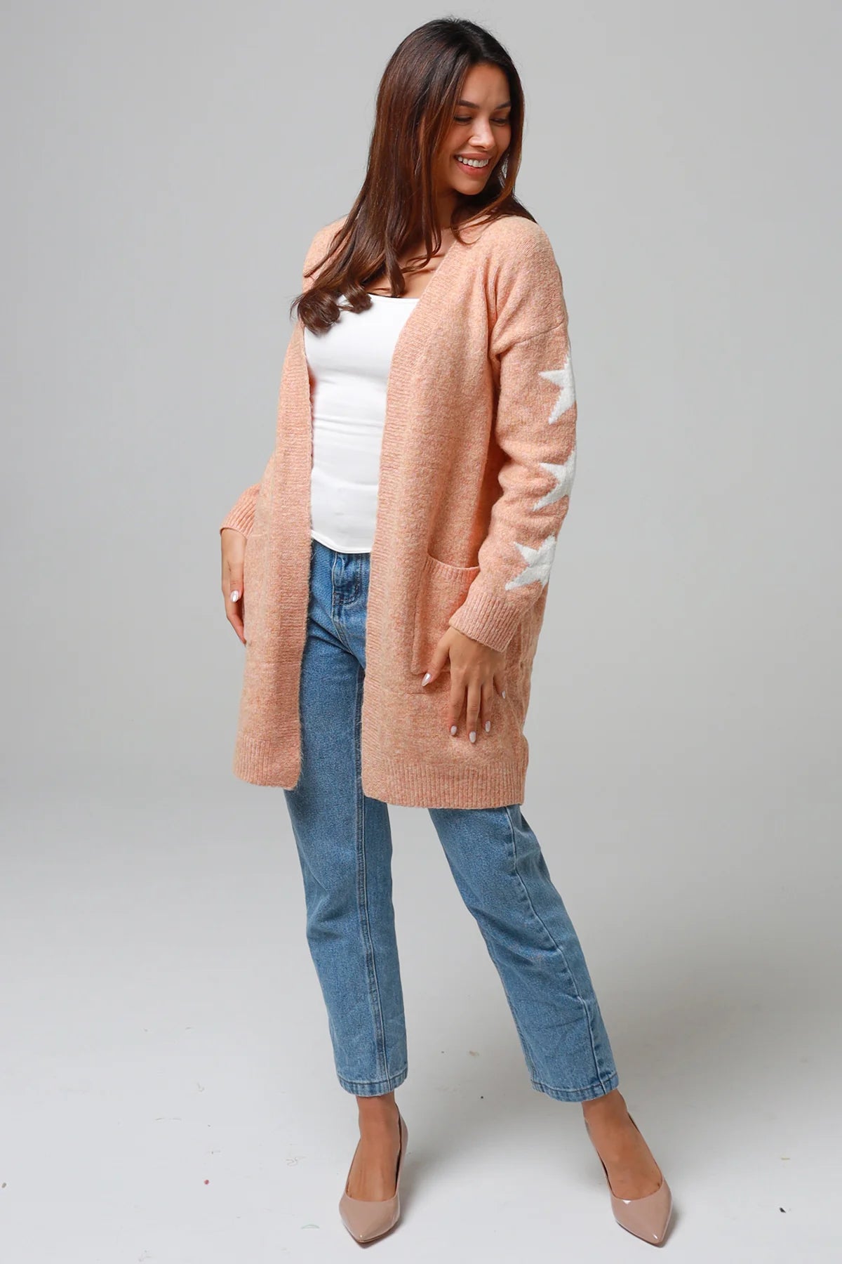 Woman wearing a peachy-blush longline cardigan with white star accents, front pockets, and relaxed fit — cozy knitwear styled with jeans and a white top, perfect for layering.