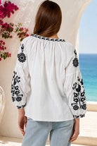 A woman viewed from behind wearing a white blouse with black floral embroidery on the sleeves and neckline, paired with light blue jeans, set against a Mediterranean-style background with a blue sea and white stucco walls.