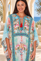 A woman modeling a long, flowing kaftan dress with a colorful floral and botanical print adorned with sparkling rhinestones standing on a sunlit outdoor patio with a view of the ocean in the background | Bohemian Love Runway 

