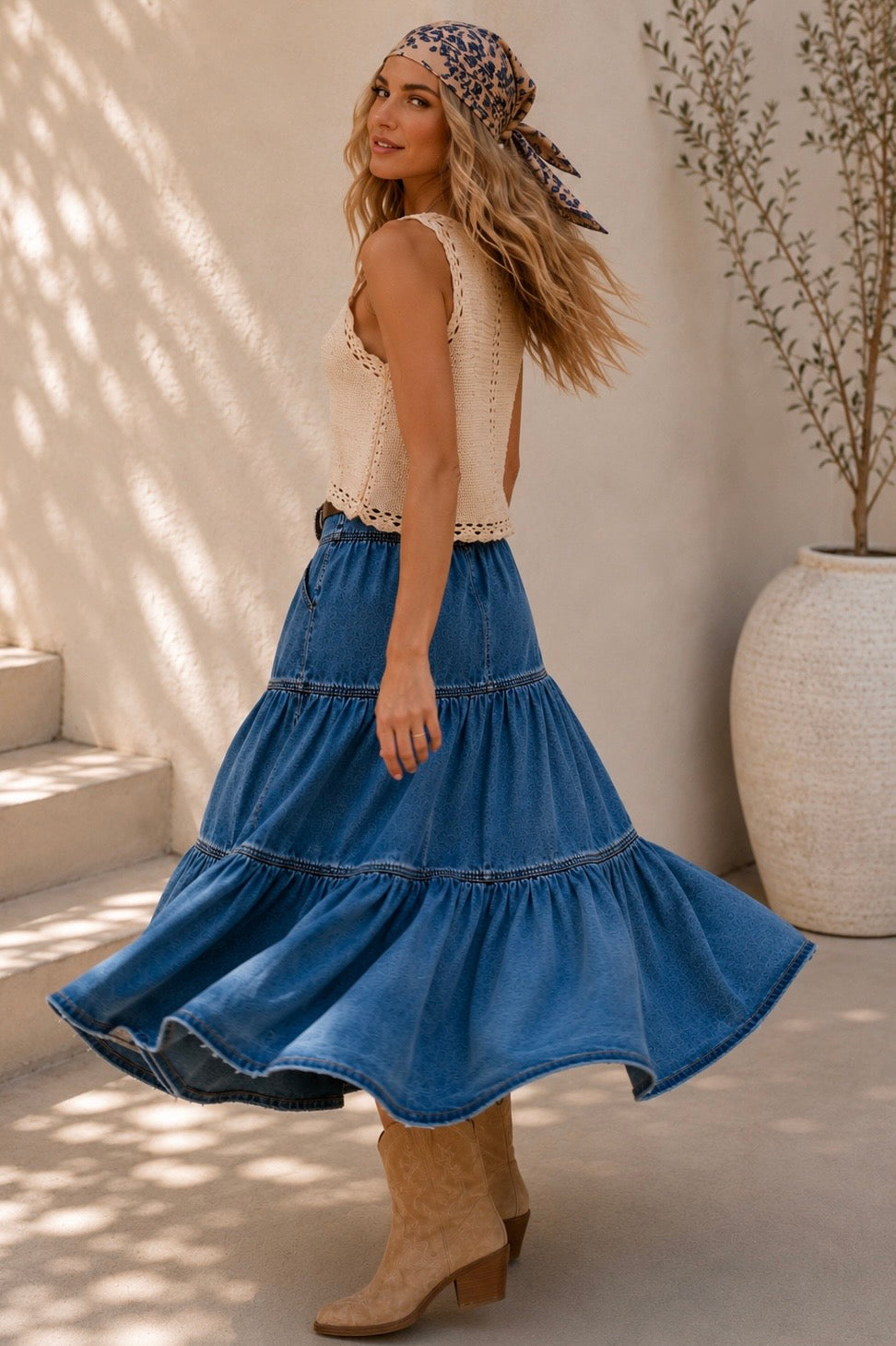 A woman models a bohemian-style outfit featuring a high-waisted, tiered blue denim skirt with a decorative belt, paired with a cream crochet top and beige boots, set against a sunlit neutral background.