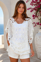 A woman wearing a white eyelet lace cover-up top with cutout details, standing outdoors in front of a white stucco wall with a blue ocean and pink flowers in the background.