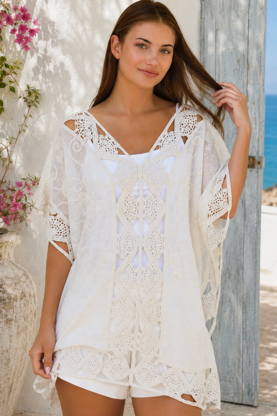 Cream crochet boho kaftan with ornate lace panels and relaxed sleeves in a sunlit Mediterranean coastal setting.
