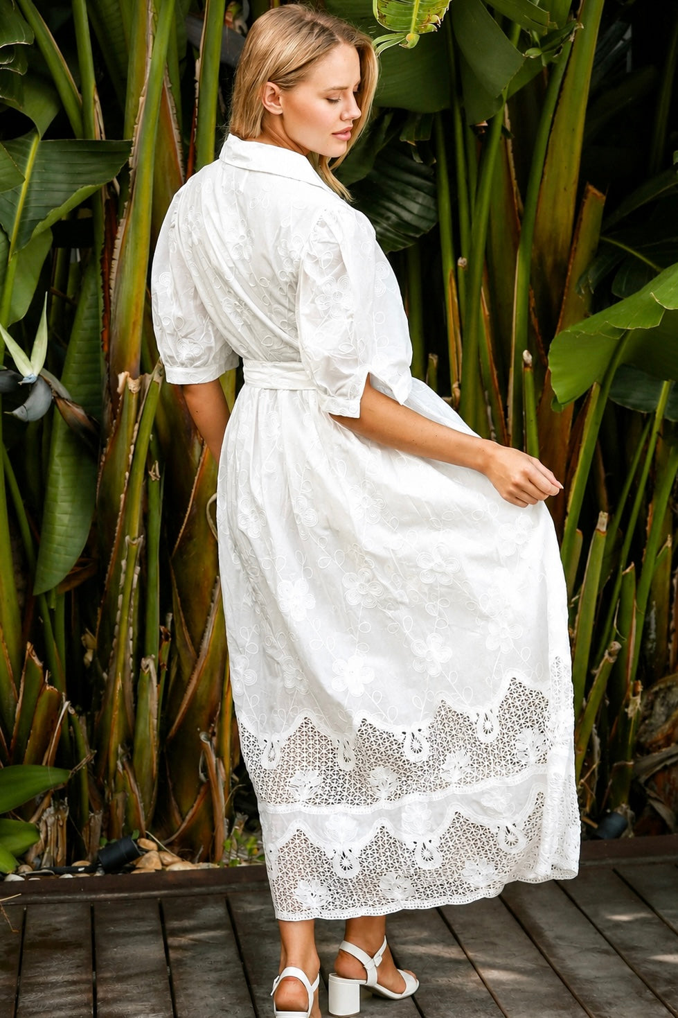 A woman wearing a white lace midi shirt dress with short puff sleeves, a tie waist, and a front slit, standing outdoors in front of tropical green foliage.