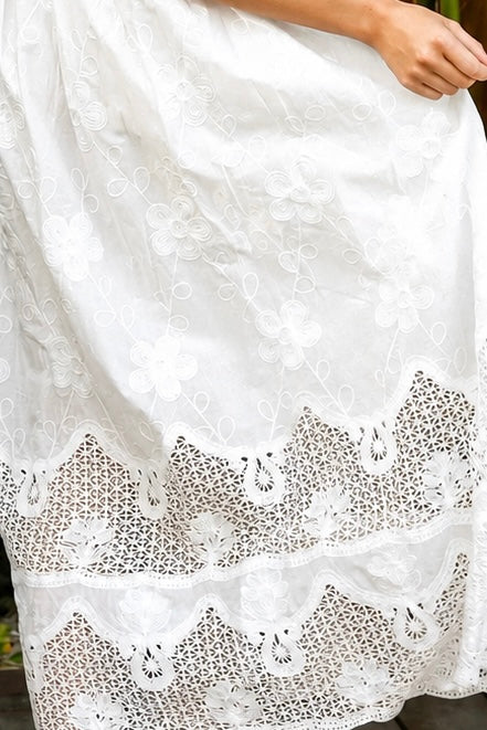 A woman wearing a white lace midi shirt dress with short puff sleeves, a tie waist, and a front slit, standing outdoors in front of tropical green foliage.