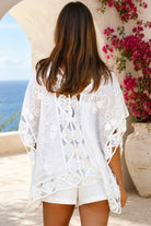 A woman wearing a white eyelet lace cover-up dress with cutout details, standing outdoors in front of a white stucco wall with a blue ocean and pink flowers in the background.