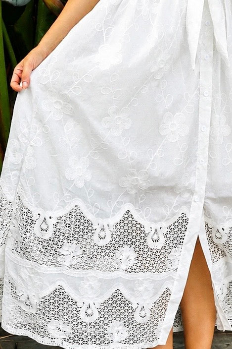 A woman wearing a white lace midi shirt dress with short puff sleeves, a tie waist, and a front slit, standing outdoors in front of tropical green foliage.