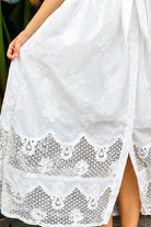 A woman wearing a white lace midi shirt dress with short puff sleeves, a tie waist, and a front slit, standing outdoors in front of tropical green foliage.