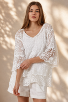 A woman wearing a white lace long-sleeve tunic top with matching white shorts, standing in a sunlit outdoor setting with a beige wall and potted plant in the background.