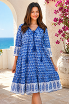 A woman wearing a blue and white patterned bohemian-style dress, standing in front of a Mediterranean-style stone wall with a view of the blue sea in the background from Bohemian Love Runway 