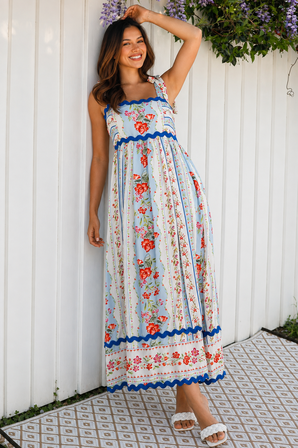 A woman wearing a sleeveless, A-line maxi dress with a white base, blue scalloped trim, and a red and pink floral print, standing outdoors in front of green plants.